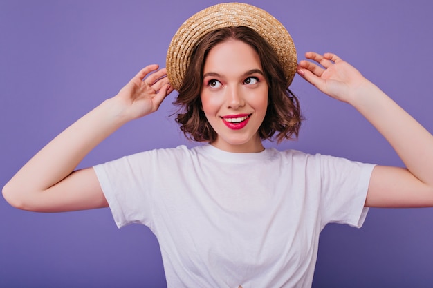 紫色の壁に笑顔でポーズをとる白いTシャツのデボネア暗い目の女の子。麦わら帽子に触れている熱狂的な白人女性の屋内写真。