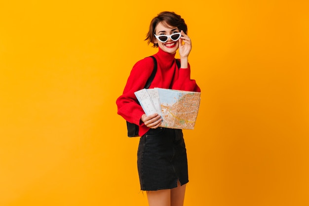 Debonair female tourist touching sunglasses on yellow wall