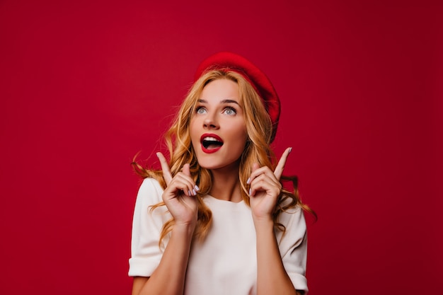 Debonair french girl posing on red wall. Dreamy girl in elegant beret expressing sincere emotions.