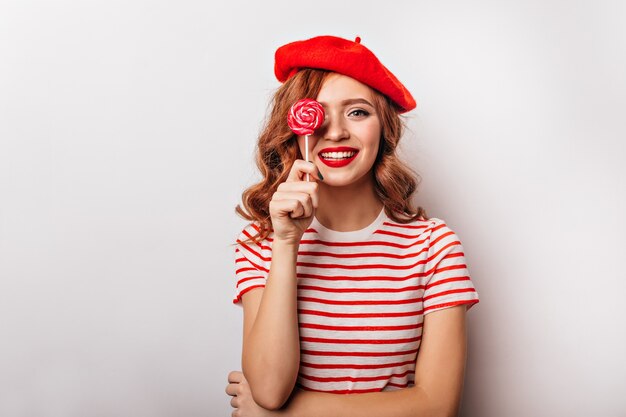 Debonair girl in red beret posing with lollipop. Dreamy french female model standing on white wall with candy.