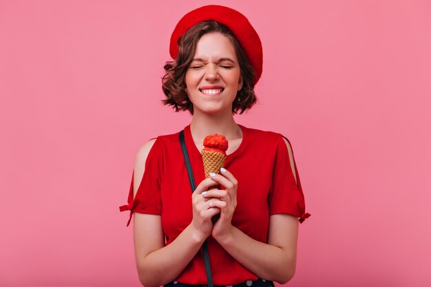 Debonair girl with short haircut eating ice cream with pleasure. Indoor photo of cheerful white lady in red clothes standing.