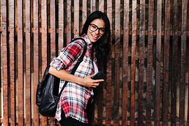 Debonair tanned lady in glasses having fun during photoshoot. Outdoor photo of magnificent girl with trendy backpack standing on wooden wall.