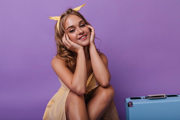 Бесплатное фото Обаятельная загорелая женщина сидит рядом с чемоданом студийный снимок улыбающейся заинтересованной блондинки, готовящейся к отпуску