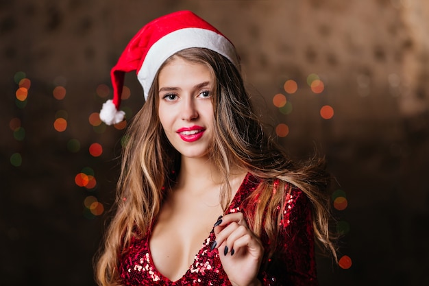 Debonair woman in santa claus hat posing on brown wall