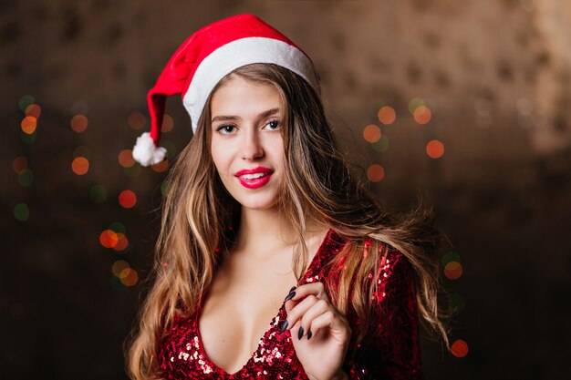 Debonair woman in santa claus hat posing on brown wall