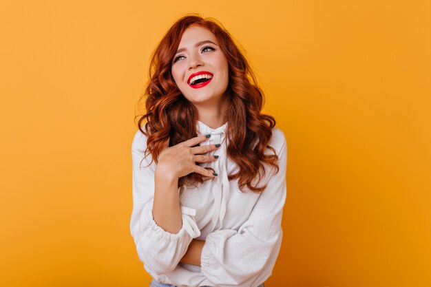 Debonair woman with wavy hairstyle laughing on yellow wall. Indoor photo of graceful ginger lady in white blouse.