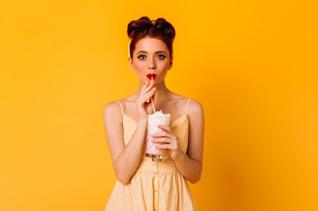 Debonair young lady drinking milkshake. Beautiful red-haired girl in pinup attire standing on yellow space.