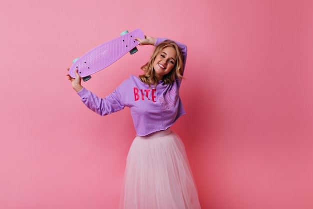 Debonair young lady in lush skirt chilling. Graceful fair-haired girl holding pink skateboard.