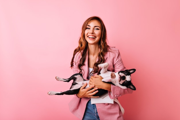 Debonair young woman in stylish jacket holdng funny french bulldog. portrait of jocund long-haired girl spending time with her dog.