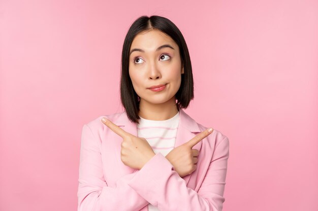 Decision concept Young asian businesswoman corporate worker pointing fingers sidways two directions showing variants looking indecisive while choosing pink background