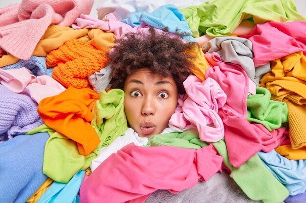 Decluttering second hand spring cleaning fast fashion and organization of life. Stunned Afro American woman with curly hair looks through big heap of colorful clothes puts things in order at closet