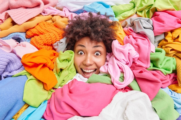 Foto gratuita concetto di decluttering e riordino. la donna afroamericana riccia positiva impressionata ordina i vestiti nell'armadio coperto con una pila di vestiti multicolori organizza l'armadio rivende gli abiti usati