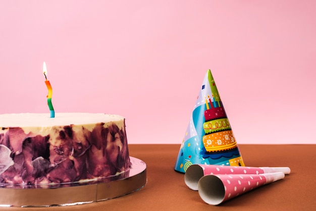 Decorative birthday cake with party hat and horn blowers on desk against pink background