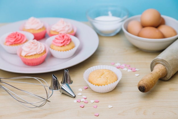 Decorative cupcake with ingredients; whisk; sprinkle; rolling pin and nozzles on wooden desk