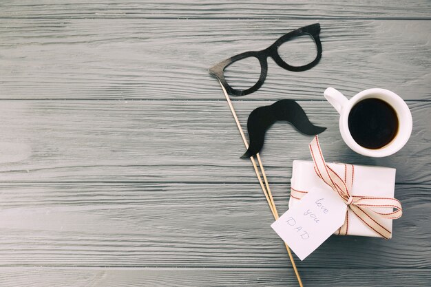 Decorative eyeglasses and moustache near present with mug of beverage