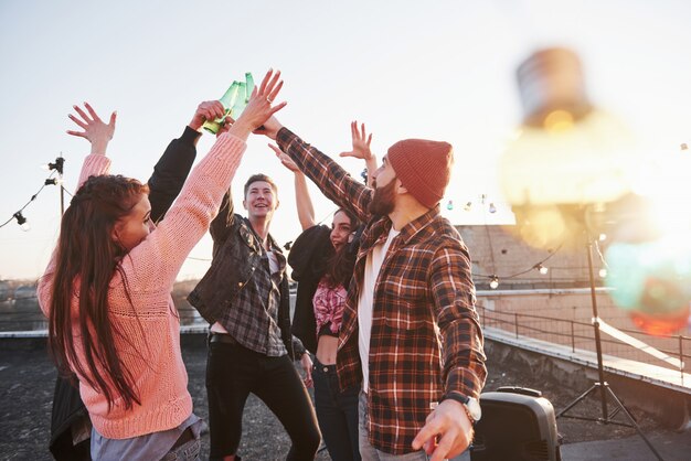 Decorative festive light bulbs. Holidays on the rooftop. Cheerful group of friends raised their hands up with alcohol