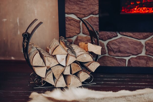 Decorative metallic holder with heap of wooden logs next to stony fireplace with burning logs.