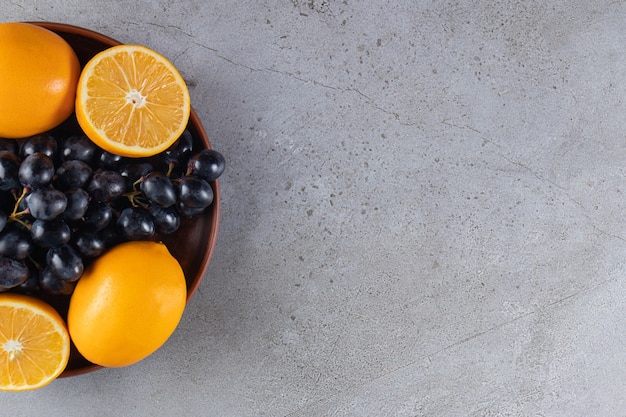 Deep plate of fresh black grapes and oranges on stone table.