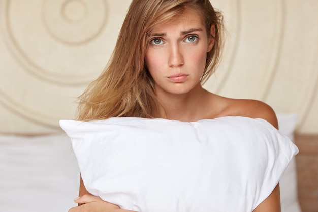 Free photo deeply upset young woman sits on bed at home, feels lonely and sad, suffers from insomnia, embraces pillow or has some disagreement with boyfriend after spending night together. sleepless concept