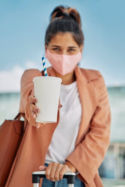Defocused woman with medical mask holding drink and luggage and at the airport during pandemic