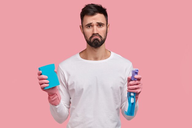 Dejected gloomy man with dark stubble, frowns face in displeasure, holds washing spray and sponge, cleans room alone, has fatigue look