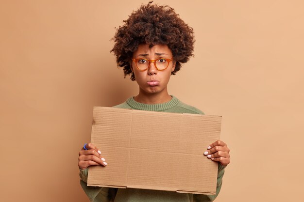 Удрученная грустная женщина поджимает губы и печально смотрит вперед, держит пустой картон для вашего рекламного контента, носит оптические очки, изолированные на бежевой стене
