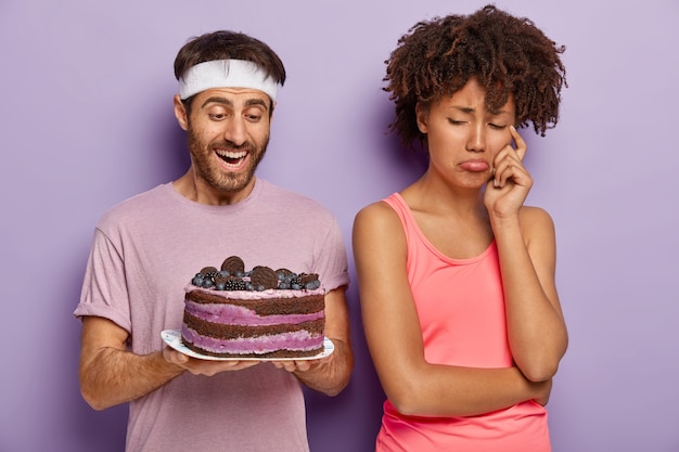 Dejected upset woman turns from husband who holds tasty cake on plate, has sad expression as cannot eat sweet desserts for keeping fit and slim leads healthy lifestyle, refuses eating junk food