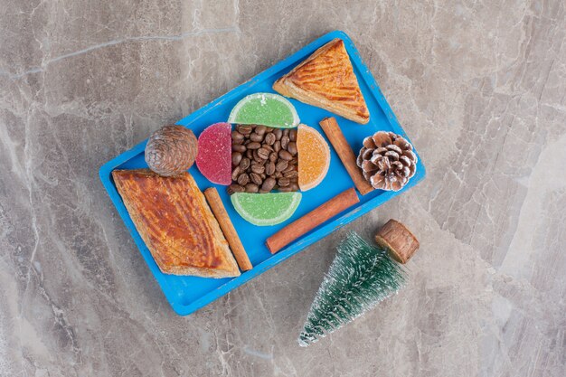 Delectable arrangement of marmelade, coffee beans, kyatas, cinnamon sticks, pine cones and a tree figurine on marble. 