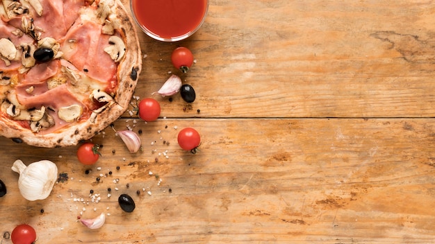 Foto gratuita pancetta deliziosa; pizza ai funghi vicino all'aglio; pomodoro ciliegino; oliva nera e ciotola di salsa di pomodoro sulla scrivania in legno