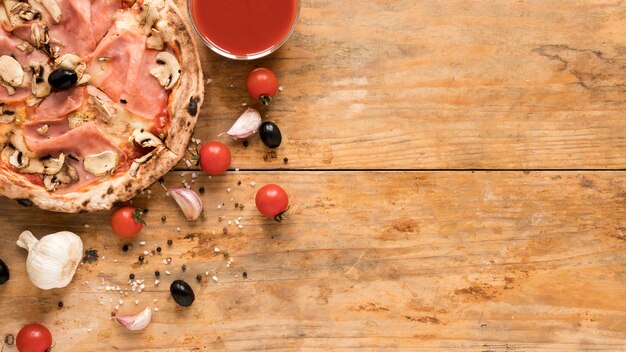 Delicious bacon; mushroom pizza near garlic; cherry tomato; black olive and bowl of tomato sauce over wooden desk