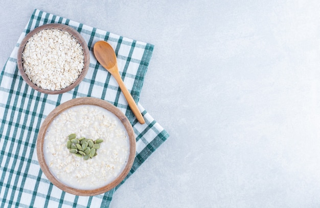 Delicious breakfast serving of pepitas-topped oatmeal on folded tablecloth, next to a bowl of oat and a wooden spoon on marble background.