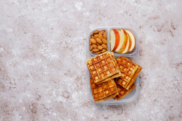 Delicious breakfast with almonds,red apples slices, waffles,croissants on plastic lunch box on light 