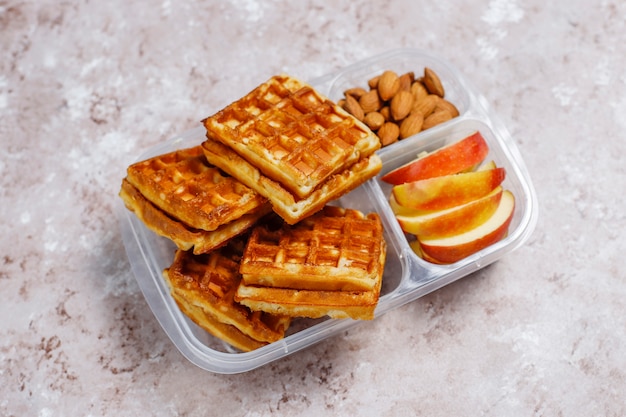 Delicious breakfast with almonds,red apples slices, waffles,croissants on plastic lunch box on light 