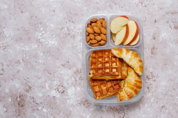 Delicious breakfast with almonds,red apples slices, waffles,croissants on plastic lunch box on light 