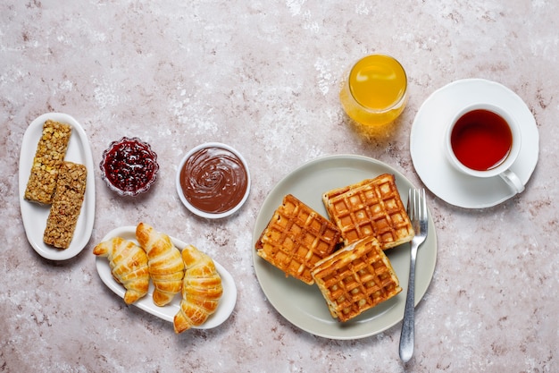 Вкусный завтрак с кофе, апельсиновым соком, вафлями, круассанами, джемом, ореховой пастой на свету, вид сверху