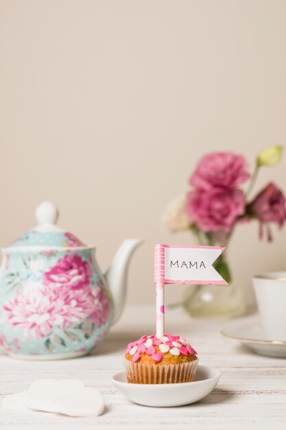 Вкусный торт с декоративным флагом с названием мамы возле чайника и цветов