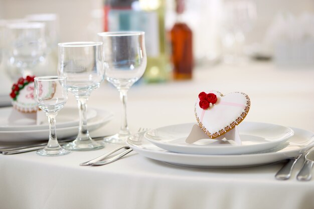 Delicious cookie covered with white sweet glaze and decorated with little red roses and white tiny pearls stands on table, served with wine glasses. Good decoration for for festive wedding table.