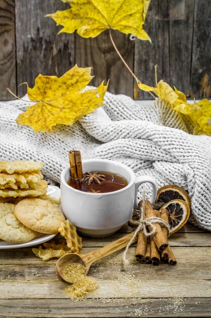 вкусное печенье и чашка горячего чая с палочкой корицы и ложкой коричневого сахара на дереве с желтыми осенними листьями,
