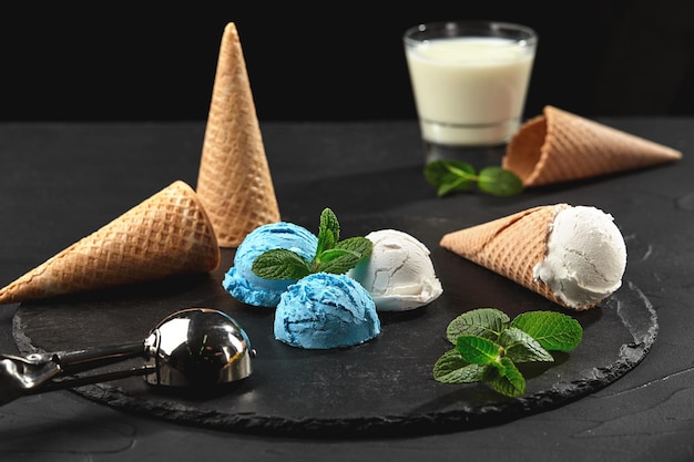 Delicious creamy and blueberry ice cream decorated with mint, and waffle cones are served on a stone slate, standing on a dark table over a black background. Metal scoop and a glass of milk is nearby.