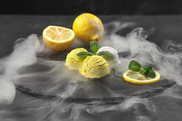 Вкусное сливочное и цитрусово-лимонное мороженое, украшенное мятой и подаваемое в дыму на каменном сланце на черном фоне. Снимок крупным планом.