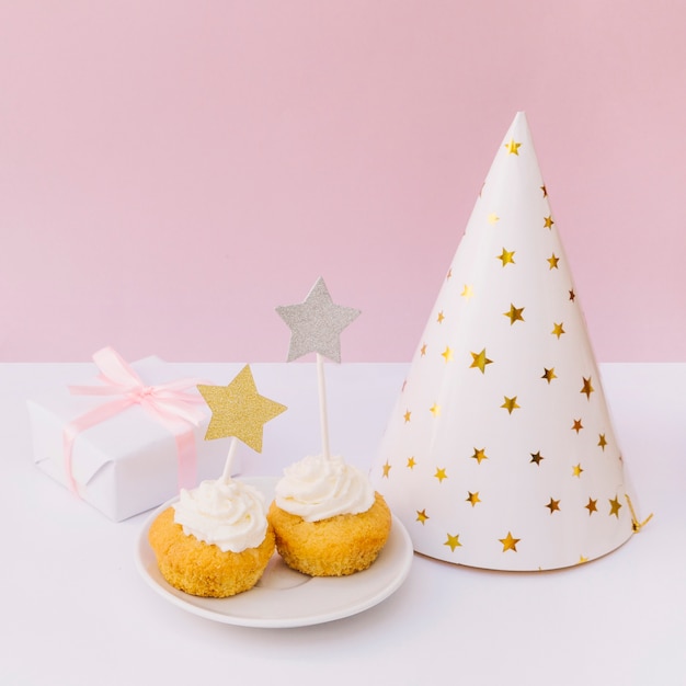 Вкусный кекс; шляпа для вечеринок; подарочная коробка и кексы на белой деревянной поверхности на розовом фоне