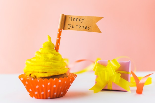 Delicious cupcake with happy birthday flag near gift on white surface