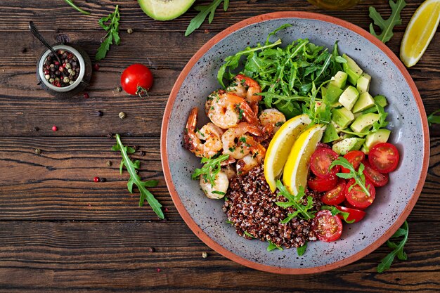 Delicious healthy Buddha bowl with shrimps, tomato, avocado, quinoa, lemon and arugula on the wooden table. Healthy food. Top view. Flat lay.