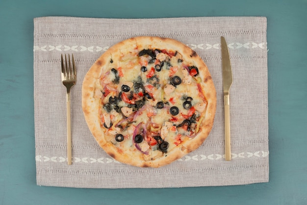 Вкусная горячая пицца с оливками и помидорами на синем столе с золотыми столовыми приборами.