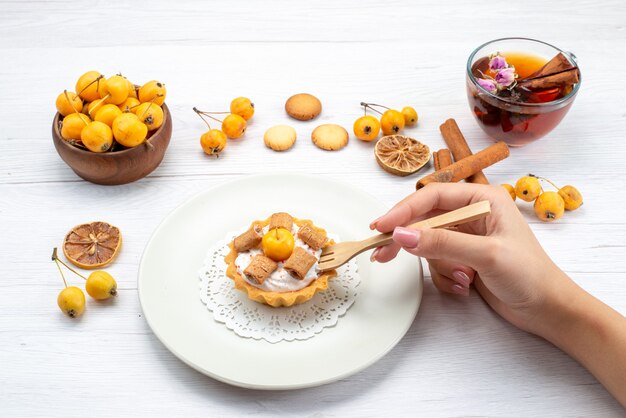 delicious little cake getting eat by female along with yellow cherries cinnamon coookies and tea on light desk, cookie cake biscuit sweet