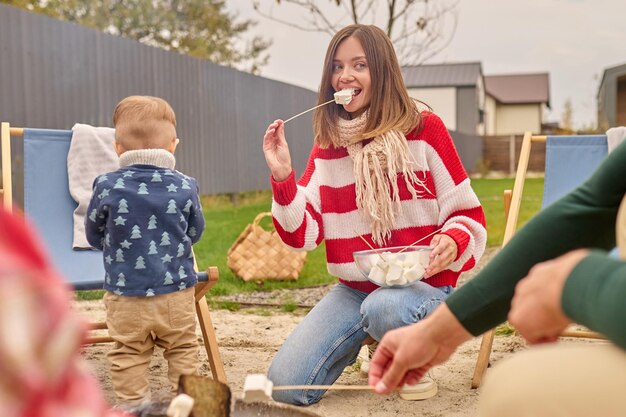 Delicious marshmallows. Young pretty woman with long blond hair in striped sweater crouching holding plate biting off marshmallows looking aside and husband with children near campfire outdoors