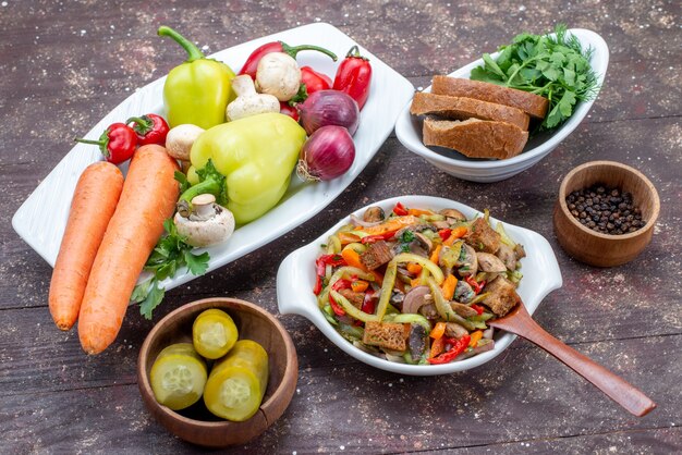 вкусный мясной салат с нарезанным мясом и приготовленными овощами вместе с солеными огурцами, хлеб, зелень на коричневом столе, еда, блюдо, мясо