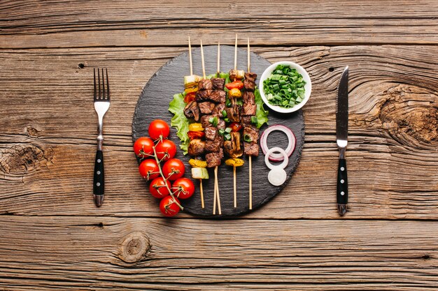 Delicious meat skewer on black slate with fork and butter knife over wooden table