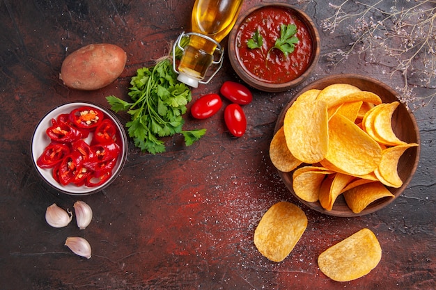 Free photo delicious potato crispy chips in a small brown bowl fallen oil bottle green tomatoes garlic ketchup chopped pepper on dark table