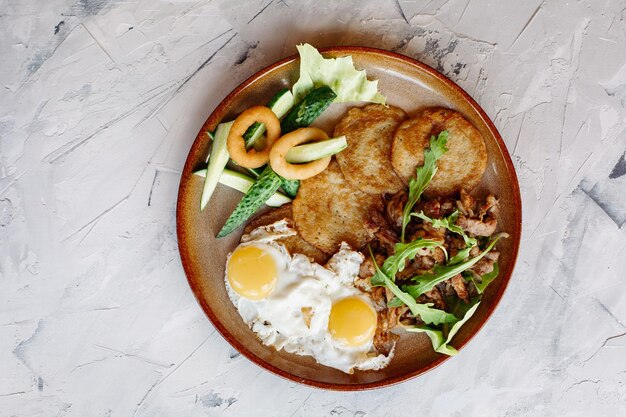 Delicious potato puncakes served with fried eggs laying on glancy clay plate including fresh green cucumbers fried golden onion and crusty meat served with salad leaves Looking tasty White table